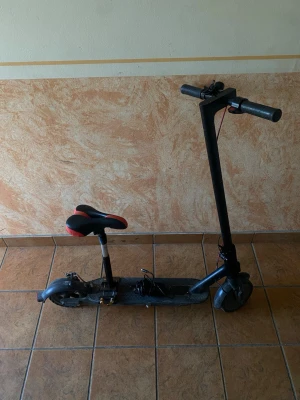 Svart elsparkcykel med sadel - Elektrisk sparkcykel i svart med sadel som har röda detaljer. Utrustad med laddare och bred fotplatta. Styret är rakt och cykeln har två hjul. Perfekt för smidig transport i stan eller till skolan.