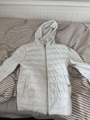 HM cardigan - Ljusgrå pufferjacka med quiltad design och dragkedja framtill. Jackan har en stor huva med dragsko och två fickor med knappstängning. Tillverkad i ett mjukt material som ger en avslappnad och modern look.