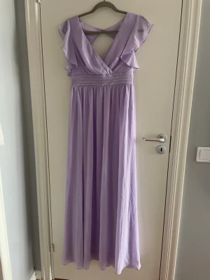 Ljuslila långklänning med volang - Säljer en elegant ljuslila långklänning med v-ringning och volangdetaljer vid axlarna. Klänningen har ett markerat midjeband och ett luftigt, svepande fall. Perfekt för dig som vill ha en romantisk och stilren look. Den har lappen kvar för jag kunde tyvär inte ha den. Prisen går att diskutera 💘