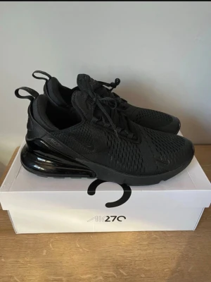 Nike Air Max 270 helsvarta  Finns även i 42-43 - Snygga helsvarta Nike Air Max 270 sneakers med mesh-ovandel och stor synlig Air-enhet i hälen. Skorna har en sportig siluett, svarta skosnören och diskreta Nike-loggor på sidan. Perfekta för dig som gillar stilrena och bekväma sneakers.
