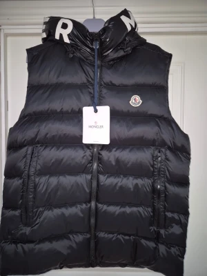 Svart dunväst från Moncler - Snygg svart dunväst från Moncler med huva och dragkedja framtill. Västen har två stora fickor med dragkedja och Moncler-logga på bröstet. Puffig design och detaljerad dragkedja med Moncler-märke. Perfekt för lager-på-lager och streetwear.