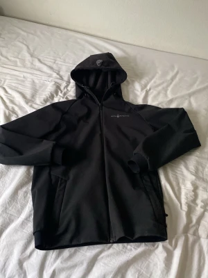 Svart vindjacka från Sail Racing - Snygg svart vindjacka från Sail Racing med huva och hel dragkedja framtill. Jackan har två sidofickor med dragkedja och diskret logga på bröstet. Tillverkad i ett mjukt och smidigt material som skyddar mot vind. Perfekt för dig som gillar stilren och sportig look.