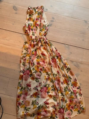 Blommig långklänning med knytband - Långklänning i ljus bas med blommigt mönster i rosa, gult och grönt. Klänningen har halterneck, markerad midja med knytband och ett luftigt, svepande fall. Perfekt för sommarens alla tillfällen.