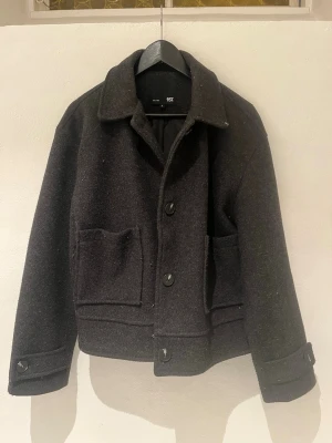 Mörkgrå jacka  - Stilren mörkgrå ulljacka från lager157 med klassisk krage, stora framfickor och knäppning med stora knappar. Jackan har en rak, något croppad passform och långa ärmar med knappdetaljer vid mudden. Perfekt för kyliga dagar.