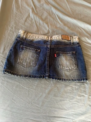 Blå jeanskjol från bens - Säljer en kort jeanskjol från bens i blå denim med slitna detaljer och rå kant. Kjolen har klassiska fem fickor, bälteshällor och dragkedja fram. Perfekt för en avslappnad och trendig look.
