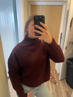 Vinröd polotröja stickad - Mysig vinröd stickad polotröja med hög krage och avslappnad passform. Tröjan har långa ärmar och ribbade muddar vid ärmslut och nederkant. Perfekt för kalla dagar och enkel att styla med jeans eller kjol.