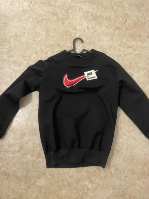 Svart sweatshirt från Nike - Cool svart sweatshirt från Nike med röd swoosh-logga och patch med texten 'Nike Sportswear' på bröstet. Tröjan har rund halsringning och ribbade muddar vid ärmslut och nederkant. Perfekt för en sportig och avslappnad stil.