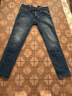 Blå raka jeans från replay  - Säljer ett par klassiska blå jeans från replay med raka ben och femficksmodell. Jeansen har snygga slitningar och en ljusare tvätt på låren. Tillverkade i slitstark denim med läderpatch bak i midjan och ikonisk bakficka med V-söm. Anbass modell.