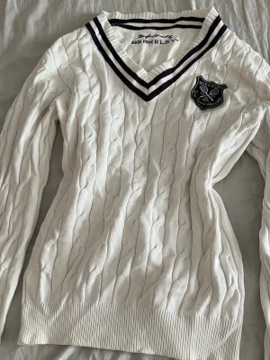 Vit kabelstickad tröja Berkeley - Kabelstickad vit tröja från Berkeley med V-ringad hals och mörklila ränder. Tröjan har ett klubbmärke på bröstet och ribbade muddar vid ärmslut och nederkant. Perfekt för en sportig och klassisk look.