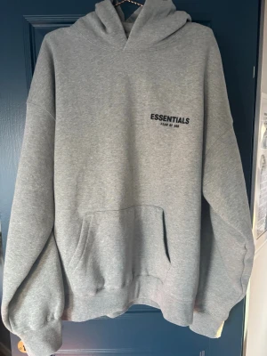 Grå Essentials Fear of God hoodie - Snygg grå hoodie från Essentials Fear of God med svart logga på bröstet och stor text på ryggen. Klassisk känguruficka framtill, ribbade muddar och stor huva. Tillverkad i mjuk bomullsblandning som är perfekt för en avslappnad streetwear-look.