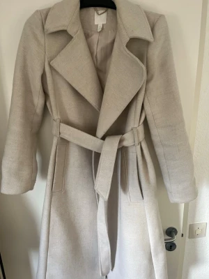 Beige kappa med knytskärp - Stilren beige kappa med bred krage och knytskärp i midjan. Kappan har långa ärmar och två sidofickor. Materialet ser ut att vara en mjuk ullblandning som passar perfekt för kyligare dagar. Enkel och tidlös design som funkar till många outfits.