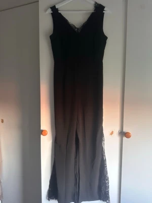 Svart jumpsuit med spetsdetaljer - Snygg svart jumpsuit med spetsdetaljer vid halsringning och längs bensluten. Ärmlös modell med v-ringning och raka ben. 