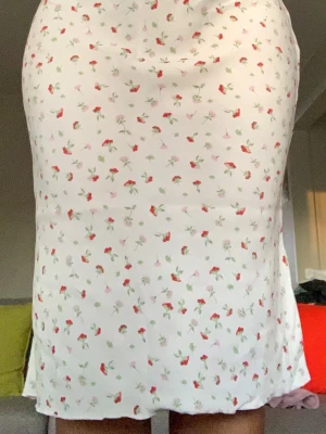 Vit blommig långkjol - Söt vit långkjol med små röda och gröna blommor. Kjolen har en rak passform och är tillverkad i ett lätt och mjukt material som känns skönt mot huden. Perfekt för varma dagar och ger en romantisk vibe till din outfit.