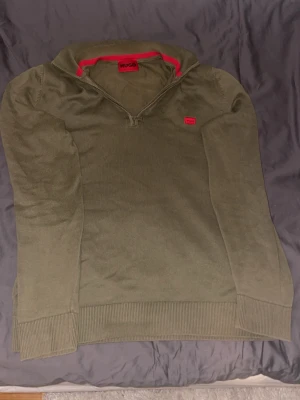 Olivgrön stickad tröja från HUGO - Snygg olivgrön stickad tröja från HUGO med halv dragkedja och ribbade muddar. Tröjan har en röd HUGO-logga på bröstet och röd detalj på insidan av kragen. Perfekt för dig som gillar stilrena plagg med en twist.