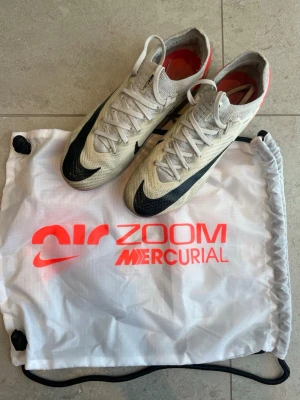 Nike Air Zoom Mercurial Vapor 15 fotbollsskor AG - Säljer ett par Nike Air Zoom Mercurial Vapor 15 fotbollsskor i vitt med svarta och röda detaljer. Skorna har en smal, aerodynamisk form och är tillverkade i lätt syntetmaterial för bästa känsla på planen. Klassisk snörning och dobbar för spel på konstgräs. Skorna köptes från original pris för 3099kr .Medföljer originalpåse.