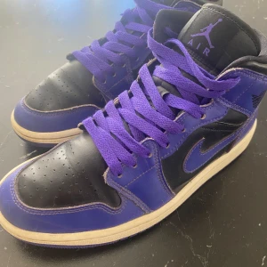 Nike Air Jordan 1 svart/lila sneakers - Säljer ett par Nike Air Jordan 1 sneakers i svart och lila skinn med klassisk Jordan-logga på sidan. Skorna har lila snören, svart tåparti och vit sula. Perfekta för dig som vill sticka ut med en ikonisk och sportig look.