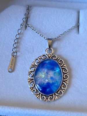 Halsband med blått ovalt självlysande hänge - Snyggt halsband i rostfritt stål med ett stort ovalt hänge i blått, lila och grönt. Efter att ha varit i vanligt dagsljus eller solljus så lyser hänget i ett starkt blått sken i mörker. Hänget har en dekorativ ram med snirkliga detaljer som ger en vintagekänsla. Kedjan är tunn och justerbar i rostfritt stål. Perfekt för dig som gillar färgstarka accessoarer. Handgjort, både kedjan och hänget tål vatten utan att missfärgas. 