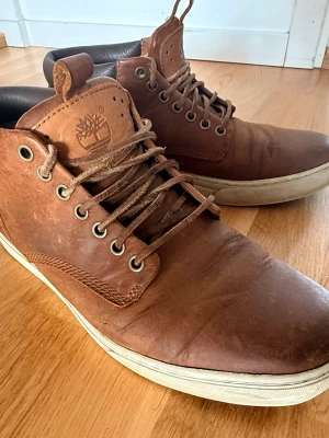 Bruna läderskor Timberland - Säljer ett par bruna läderskor från Timberland i äkta läder med vita sulor och klassisk logga på plösen. Skorna har snörning med metallhål och en mellanhög design som ger en snygg streetstil. Perfekta för dig som gillar en robust och stilren look.
