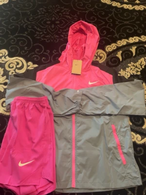 Nike träningsset rosa och grå - Säljer ett snyggt träningsset från Nike med en rosa och grå vindjacka med huva och dragkedja samt matchande rosa träningsshorts. Jackan har fickor med dragkedja och Nike-logga på bröstet, medan shortsen har logga på benet. Perfekt för löpning eller gym.