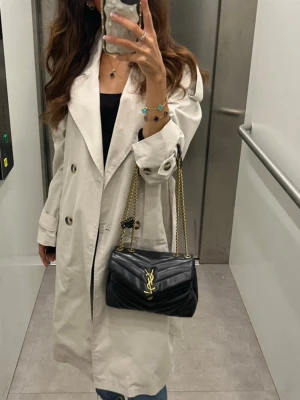 Beige trenchcoat  - Stilren beige trenchcoat med dubbelknäppning och bred krage. Klassisk modell med bälte i midjan och stora knappar framtill. Långa ärmar med justerbara remmar vid ärmsluten. Perfekt för en trendig och tidlös look.