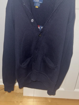 Mörkblå stickad kofta Polo Ralph Lauren - Säljer en mörkblå stickad kofta från Polo Ralph Lauren i storlek M. Koftan har v-ringning, två framfickor och knappar framtill. På ryggen finns en stor amerikansk flagga i stickat mönster med RL-broderi. Perfekt för dig som gillar klassisk och cool stil.