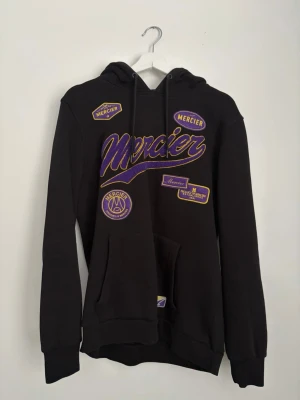 Svart hoodie från Mercier med tryck - Svart hoodie från Mercier med lila och gula broderade och tryckta patches framtill. Klassisk huva med dragsko och stor magficka. Hoodien har ribbade muddar vid ärmslut och nederkant. Perfekt för dig som gillar streetwear och vill sticka ut.