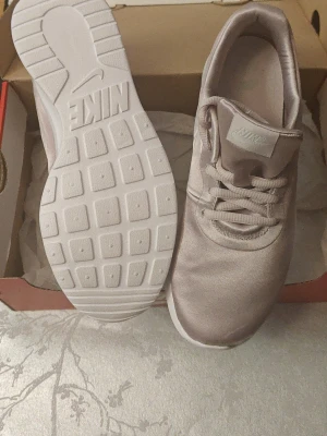 Nike ljusrosa/ beige satin sneakers 37,5 - Snygga ljusrosa/blekRosa/ beige sneakers från Nike i satinliknande material med vita sulor och klassisk Nike-logga på plösen. Skorna har låga skaft och matchande beige skosnören. Perfekta för dig som vill ha en clean och stilren look till vardagsoutfiten.Helt nya storlek 37.5 cm 23.5