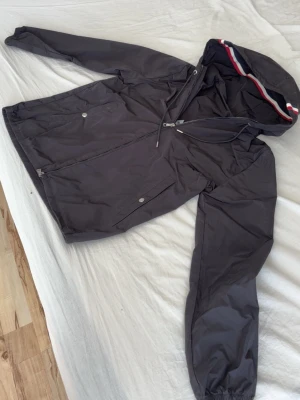 Moncler windbraker  - Snygg svart vindjacka från Moncler med dragkedja, två framfickor och huva med röd, vit och blå rand. Jackan har Moncler logga på ärmen och elastiska muddar vid ärmsluten. Perfekt nu till våren/sommaren. Har du frågor tveka inte på att höra av dig. Pris kan diskuteras 