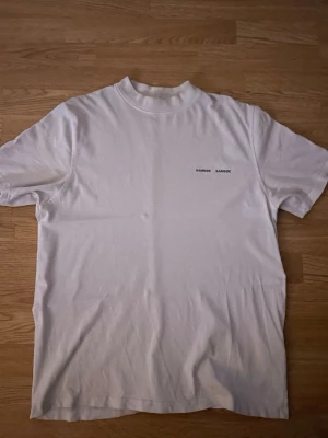 Vit t-shirt från Samsøe Samsøe - Vit t-shirt från Samsøe Samsøe med minimalistiskt tryck på bröstet. Klassisk rund hals och korta ärmar. Tillverkad i mjuk bomull för en skön och avslappnad känsla. Perfekt basplagg till garderoben. Har en liten slitning mitt på bröstet.