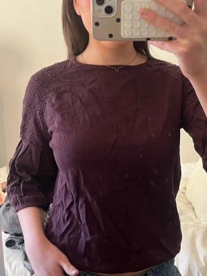 Vinröd topp med trekvartsärm - Snygg vinröd topp med trekvartsärm och strukturdetaljer på axlarna. Toppen har en rund halsringning och är tillverkad i ett mjukt material som känns skönt mot huden. 
