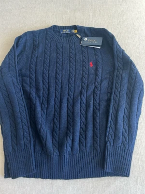 Mörkblå kabelstickad tröja Polo Ralph Lauren - Kabelstickad tröja i mörkblått från Polo Ralph Lauren med rund halsringning och ribbade muddar. Klassisk röd logga broderad på bröstet. Tillverkad i mjukt material som passar perfekt till höst och vinter.