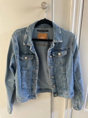 Ljusblå jeansjacka från ONLY - Klassisk ljusblå jeansjacka från ONLY med raka sömmar, två bröstfickor med knappar och silverfärgade detaljer. Jackan har en avslappnad passform och tidlös design som passar till det mesta. Perfekt att slänga över en hoodie eller t-shirt.