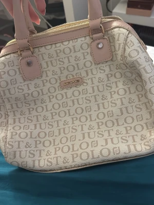 Beige handväska från Just Polo - Snygg beige handväska från Just Polo med logotypmönster över hela väskan. Väskan har ljusrosa detaljer, dubbla handtag och guldfärgade dragkedjor. Perfekt för dig som gillar stilrena accessoarer med en touch av lyx.