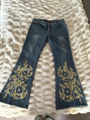 Blå lågmedjade utsvängda jeans med gula spetsdetaljer - Unika blå jeans lowrise  med utsvängda ben och stora blommiga spetsapplikationer i gult på båda benen. Nästan aldrig använda. Pris kan diskuteras💞