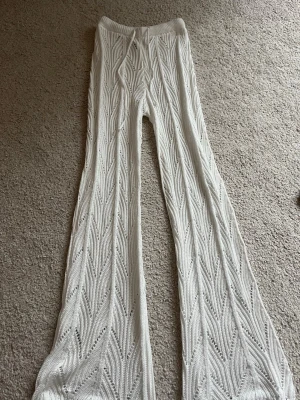 Vita virkade utsvängda leggings - Säljer ett par vita virkade leggings med snyggt mönster och utsvängda ben. De har bred resår i midjan och är genombrutna vilket ger en cool boho-vibe. Perfekta att styla över shorts eller baddräkt för en trendig look.