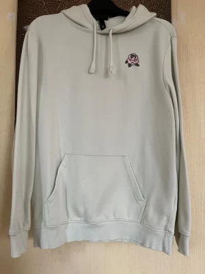 Vit hoodie med rosbrodyr från H&M - Snygg vit hoodie från H&M med regular fit och broderad rosa ros på bröstet. Hoodien har dragsko i huvan och en stor känguruficka framtill. Tillverkad i mjukt material som är skönt mot huden. Perfekt för en chill och stilren look.
