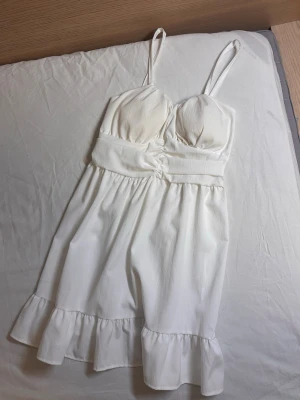 Vit kort klänning med volang - Söt vit kort klänning med smala axelband och markerad midja. Klänningen har volangkant nertill och lätt rynkade kupor för en feminin look. Perfekt för varma dagar och somriga tillfällen.