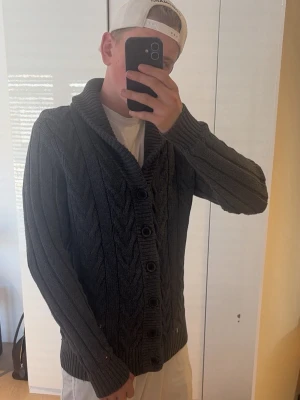 Tommy Hilfiger Cardigan - Mörkgrå kabelstickad kofta med stora knappar och sjalkrage. Koftan har långa ärmar och ribbstickade muddar vid ärmslut och nederkant. Perfekt för lager-på-lager och en chill stil.