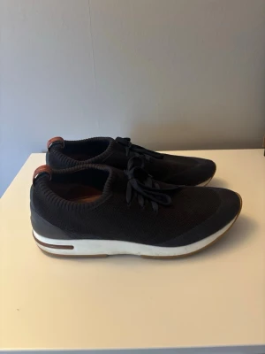 Loro Piana  - Snygga mörkblå tofflor från Loro Piana med stickad ovandel och snörning framtill. Skorna har vit sula med bruna detaljer och öppen häl för enkel påtagning. Insidan är i brunt läder för extra komfort. Perfekta för en avslappnad och trendig look. Finns en liten skråma på tån inget som märks vid användning.