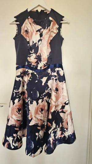 Blommig klänning med marinblå detaljer - Snygg ärmlös klänning med marinblå paneler och blommigt mönster i rosa, beige och vitt. Klänningen har markerad midja med ett satinband och utsvängd kjol. Perfekt för dig som gillar en feminin och modern stil. Använd endast 1 gång. Bandet runt midjan går att ta bort, har sytt ditt den själv.