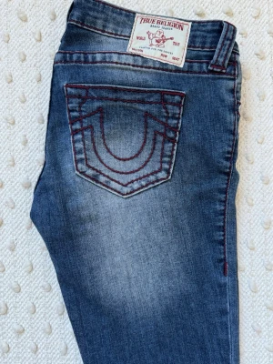 True Religion blå jeans med röd söm - Snygga blå jeans från True Religion med ikonisk röd kontrastsöm och klassisk logga på bakfickan. Jeansen har fem fickor, gylf med knapp och dragkedja samt slitningar för en cool look. Perfekta för dig som gillar streetstyle och detaljer.