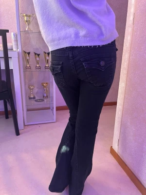 Svarta bootcut jeans med fickdetaljer - Säljer ett par svarta bootcut jeans med snygga fickdetaljer bak och knappstängning. Jeansen har låg midja och utsvängda ben, vilket ger en retro och trendig look. Perfekta för dig som vill ha en cool och avslappnad stil.