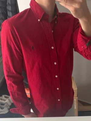 Röd skjorta från Ralph Lauren - Snygg röd skjorta från Ralph Lauren med klassisk button-down krage och broderad logga på bröstet. Skjortan har vita knappar och lång ärm, perfekt för en stilren look. Tillverkad i bomull för en skön känsla.