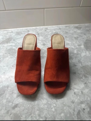 Rostbruna slip-in sandaler i mocka - Säljer ett par trendiga slip-in sandaler i rostbrun mocka med öppen tå och bred rem över foten. Sandalerna har en låg klack och en mjuk, beige innersula för extra komfort. Perfekta för dig som vill ha en stilren och enkel look.