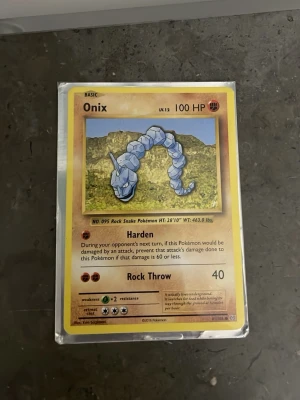 Onix Pokémonkort Base Set - Säljer ett Onix Pokémonkort från Base Set. Kortet har gul ram, Onix-illustration mot bergig bakgrund och text på engelska. Kortet är inplastat och har 100 HP samt attackerna Harden och Rock Throw. Perfekt för samlare eller Pokémonfans.