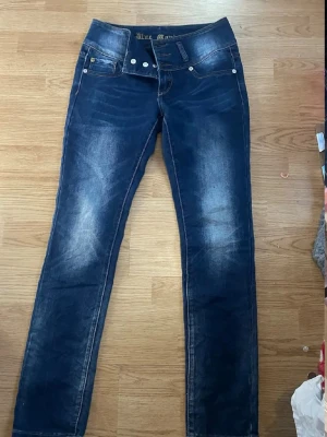 Blå skinny jeans från Blue Monkey - Snygga blå skinny jeans från Blue Monkey med slitningar och ljusare partier på låren och knäna. Klassisk femficksmodell med silverfärgade nitar och dubbla knappar i midjan. Perfekta för en trendig och avslappnad stil.