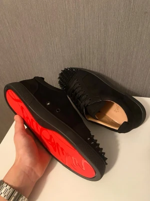 Christian Louboutin  - Säljer ett par svarta sneakers från Christian Louboutin med klassisk röd sula och svarta nitar på tån. Skorna är i mocka och har svarta skosnören. Perfekta för dig som vill sticka ut med en edgy och lyxig stil.