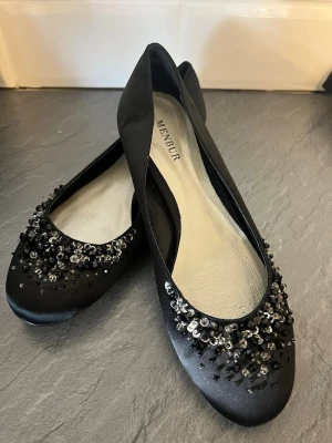 Svarta ballerinaskor med pärlor Menbur - Stilrena svarta ballerinaskor från Menbur i satinliknande material. Dekorerade med svarta och silvriga pärlor och paljetter framtill för en extra glamorös touch. Rundad tå och låg klack gör dem både snygga och bekväma.