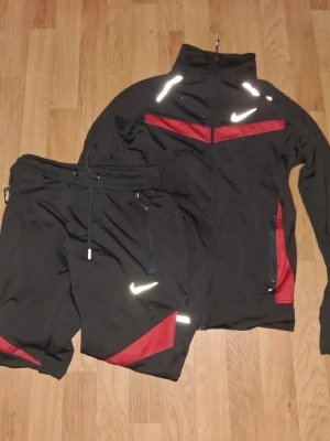 Svart Nike träningsset med röda detaljer - Säljer ett svart träningsset från Nike med röda detaljer och vita loggor. Setet består av en zip-jacka med hög krage och två fickor samt matchande byxor med dragsko i midjan. Materialet är mjukt och stretchigt, perfekt för träning eller chill.
