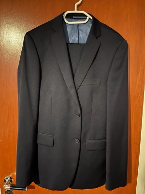 Dressmann kostymset - Snygg mörkblå kavaj från Dressmann i slim fit-modell. Kavajen har två knappar framtill, klassiska slag och fickor med lock. Fodret är ljusblått med röda detaljer. Perfekt för dig som vill ha en stilren och modern look. Kavajen har storlek 50R och kostymbyxan W30 L30.